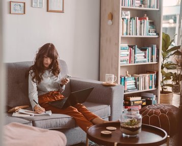 Eine Frau sitzt auf einem Sofa, nutzt einen Laptop und hält ein Smartphone in der Hand. Neben ihr liegen Unterlagen und eine Tasse auf einem Tisch. Im Hintergrund sind Bücherregale sichtbar.